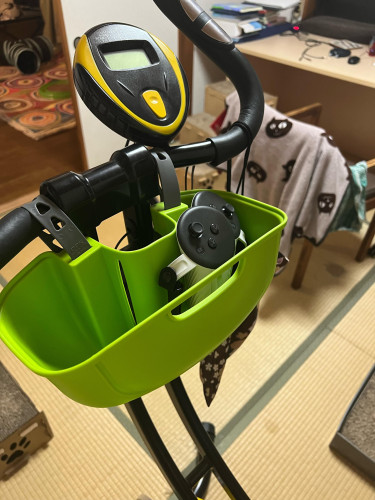 A green plastic shallow basket is strapped onto the inside of handle bars of an exercise bike. Two Quest 3 VR controllers are seen in the basket. 