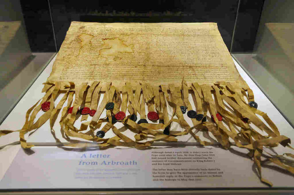 The Declaration of Arbroath. The image shows a replica of the declaration on display in a Perspex box. It is a large single sheet with closely spaced lines of writing at the end furthest from the viewer. At the near end it is cut into multiple vertical strips, each of which has a red or black wax seal on it. AT the bottom of the image is an interpretive panel giving background to the declaration.