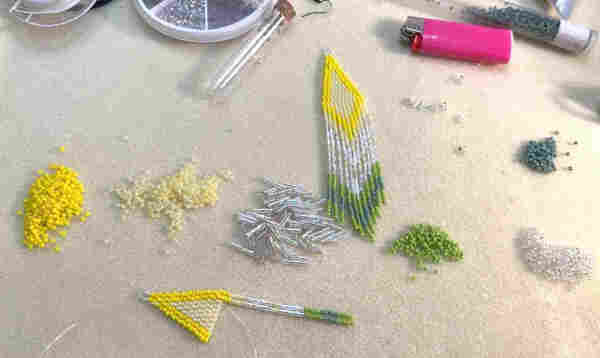 A bead mat with piles of seed beads, findings, a tube of needles, a tube of beads, and a pair of Native beaded fringe earrings in the making