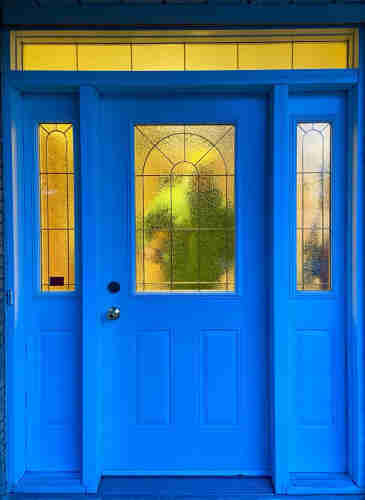 A front door of a house with decorative side panels and privacy glass. The door appears blue in the early morning light. Through the privacy glass we see yellow light, and a blurry impression of a figure wearing a green coat and yellow hat.