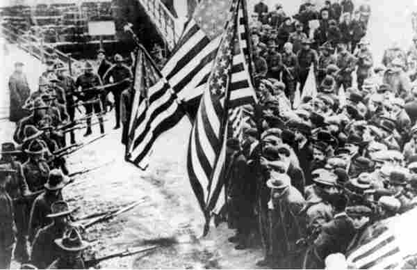 Massachusetts militiamen with fixed bayonets surround a group of strikers. Several of the strikers are carrying American flags. By http://womhist.binghamton.edu/teacher/DBQlaw2.htm, Public Domain, https://commons.wikimedia.org/w/index.php?curid=131378