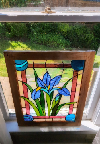 A stained glass window depicting an iris, leaning against an old craftsman-style window, looking out into a patio and grass.