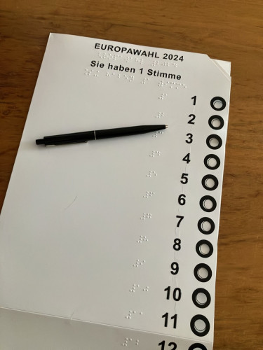 Eine Wahlschablone aus heller Pappe für die Europawahl. Darin klemmt, kaum sichtbar, ein Stimmzettel. Die Ecke rechts oben ist abgeschnitten.
In der Kopfzeile der Schablone steht in erhabenen Buchstaben "EUROPAWAHL 2024" und darunter "Sie haben 1 Stimme". Beide Texte stehen in Brailleschrift unter der Schwarzschrift. In der rechten Hälfte des Stimmzettels sind die Zahlen 1 bis 12 untereinander stehend zu sehen. Erst in Braille-, rechts davon in Schwarzschrift. Neben jeder Zahl befindet sich ein rund ausgestanztes, schwarz umrandetes  Loch, in das man sein Kreuz setzen kann. Ein schwarzer Kugelschreiber liegt quer auf der Schablone. Das Ensemble liegt auf einer Holzoberfläche.