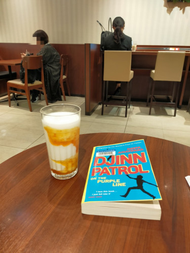 Photo is inside the cafe. On a round brown table is the turquoise paperback library book with a black illustration of a boy leaping left. On the left is a white and orange yoghurt drink in a glass. In the distance you can see 2 Japanese women sitting in the cafe, one with short hair with her face blurred and one with long hair from the back