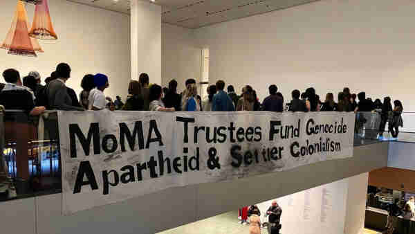 People holding a large protest banner over a railing in an indoor setting with the text "MoMA Trustees Fund Genocide Aparthied & Setter Colonialism".