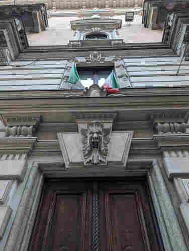 An ornate carved stone doorway with a hardwood dark brown door. Over the door is an open-mouthed, howling gargoyle. Overthat is a pair of Italian tricolor flags framing a window. More windows and stonework are visible above it.