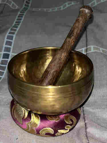 A golden colored hand beaten singing bowl sitting on a gold and purple pillow