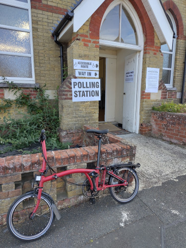 a red Brompton bike outside of a yellow brick building with temporary signs announcing it as a polling station