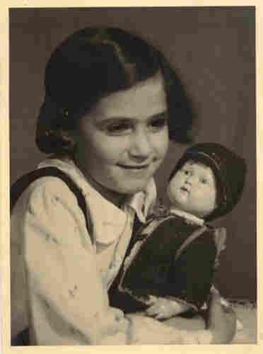 A young girl with dark hair covering her ears and a white blouse with tiny floral patterns holds a doll dressed in a jacket and cap.