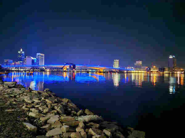 Overlooking a calm river in a late night setting along a rocky coastline with a colorful city skyline and illuminated bridge casting the numerous colorful lights upon the river's surface.