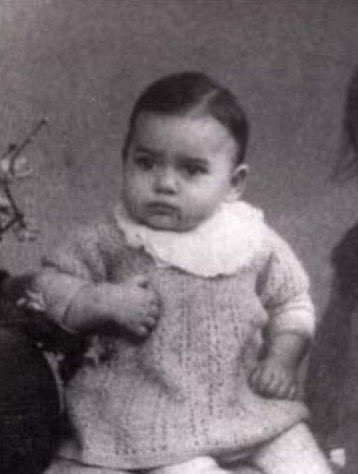 Antique black and white photo of a baby girl wearing a knitted outfit, sitting upright, with a blurred partial view of an adult in the background, suggesting a formal portrait setting.
