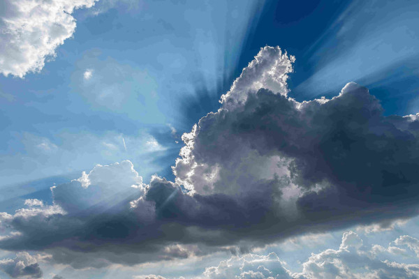 Photograph of a blue sky filled with banks of cumulus clouds. The prominent cloud bank is darkened by shadows and highlighted along the edges by the sun directly behind the clouds. The sun is very bright and it is angled in such a way that the clouds in front of it cast shadows in the sky above that is accentuated by haze that surrounds the shadows.