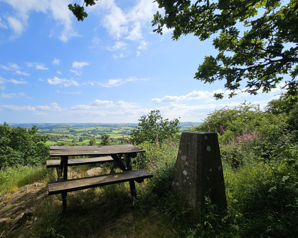 View from a hilltop across green fields. There is an old picnic bench and a concrete trig point in the foreground.