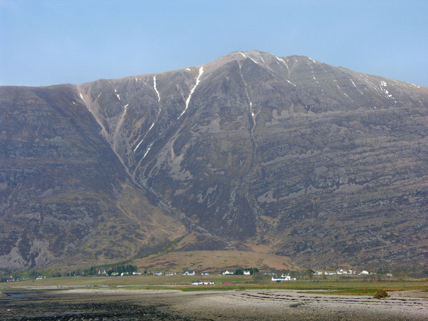 Torridon in Wester Ross. The image shows a view over stony beach in the foreground with the tide out but a little water visible on the left. Beyond it is a distant line of white buildings stretching across the frame with, above it, the huge bulk of a mountain reaching up towards the top of the frame. The summit ridge has lines of snow leading up to it in gullies. The scene is in sunlight.