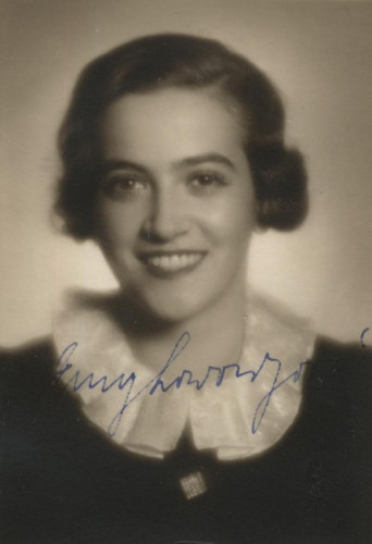 A portrait ID photo of a smiling young woman. She exposes her teeth. She has long hair styled in curls reaching behind her ears. She is wearing a dark blouse with a white semi-circular collar. 