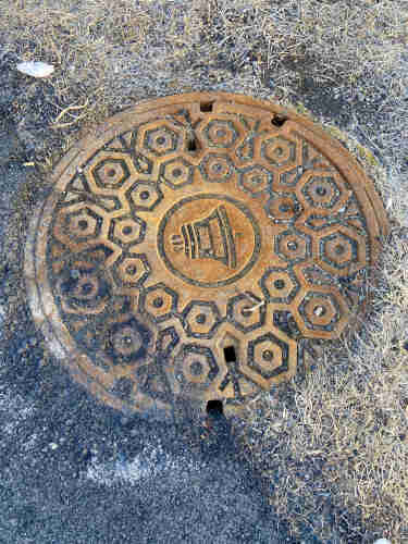 A manhole cover with a bell icon and “Bell Canada” in the center, with radiating circles of nested hexagons. The metal is rusting to an orange-brown, and the grooves of the design have filled up with dark gravel. It looks like there’s a shadow through the lower left, but it’s the gravel edge of an asphalt path that passes over the edge.