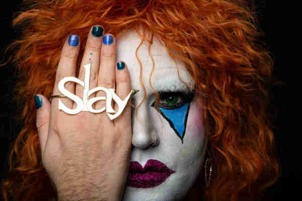 Picture of the face of a Drag queen with grren eyes. She is hiding her eye with her hand wearing a big rgin that says "Slay".