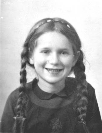 Black and white photo of a smiling child with braided hair, wearing a dark blouse. The child is looking directly at the camera, and the background is plain and unadorned. 