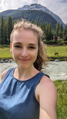 Molly taking a selfie in front of a river, with a horse in a pasture and a tall mountain behind. She's wearing a denim tank top shirt.