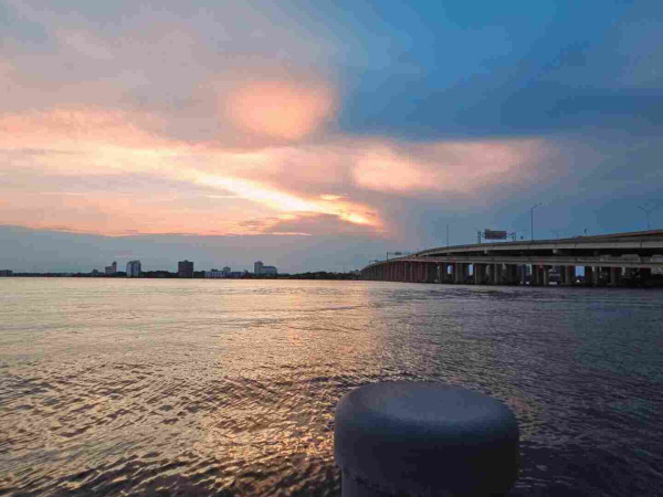 From a dock on the shoreline of a wide river, looking over the top of a grey piling where a massive interstate highway bridge crosses the river connecting the opposite shoreline with its tiny silhouetted city skyline, beneath a pink and blue sky where the sun casts colorful beams of light as it lowers in the sky, behind large, dark cloud formations.
