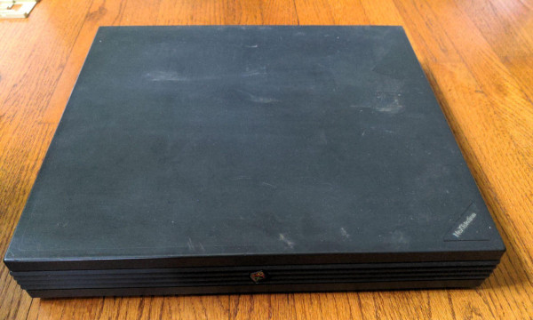 Dark gray pizza box shaped computer on a wood floor with horizontal ribs on the front and the NeXT logo in the very center.