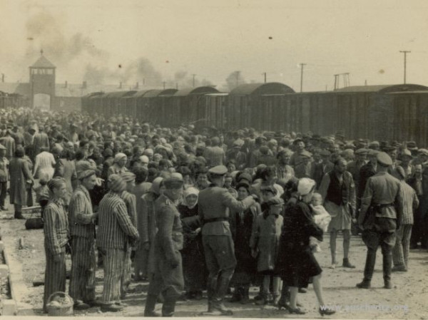 Crowds of Jews at the unloading platform at Auschwitz II-Birkenau. Some SS men and prisoners in striped uniforms visible. In the background a freight train, the gate of the camp and some smoke.