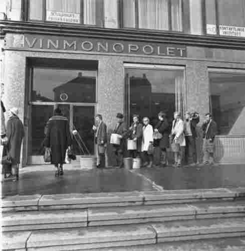 A B&W image showing people lining up outside a building with large buckets. The sign above them says "VINMONOPOLET", "The Wine Monopoly", the name of the government retailer of all alchol stronger than 4.75% by volume. This is a 1969 recreation of the result of the 1950 April Fools' joke.