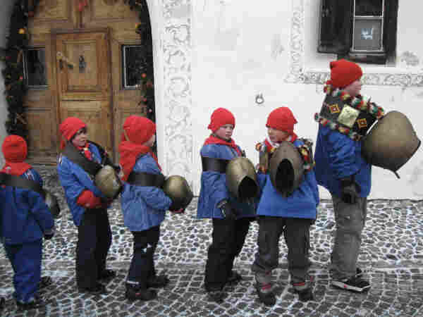 Vor einem mit Sgraffiti geschmückten Engadinerhaus haben sich sechs Kinder mit blauen Kitteln, roten Zipfelmützen und Glocken aufgereiht und warten darauf, dass der Umzug beginnt. Der Größte ganz vorne scheint seine Rolle als Anführer sehr ernst zu nehmen. Die Glocke, die er um die Schultern trägt, ist riesig.