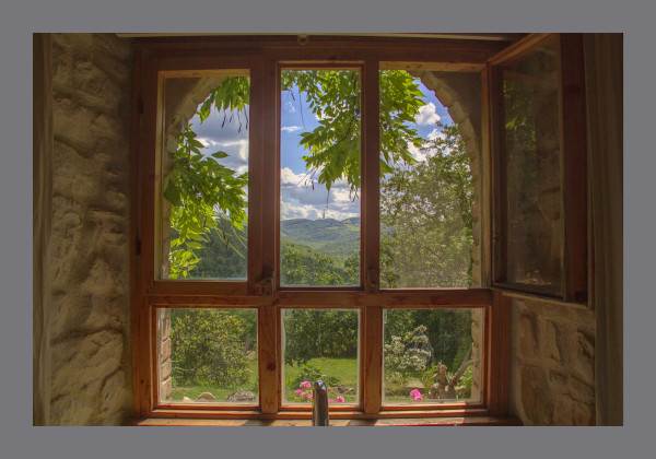 View through a 6 pane wooden window in a rough stone wall looking out to a landscape of green hills through a curtain of vegetation and pink geraniums
