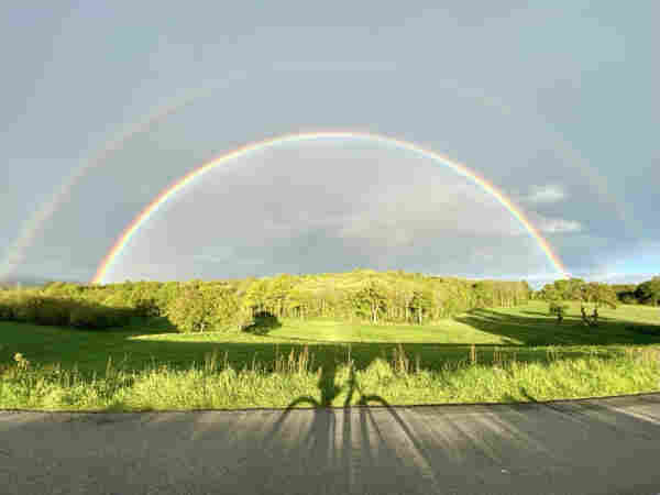 Full double rainbow over a green sunny landscape with trees, shadow of a person on a bicycle on the road in the foreground.