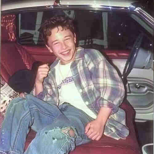 A young person seated in the backseat of a vehicle, exuding joy and laughter. The individual is casually attired in a plaid shirt layered over a graphic tee, accompanied by distinctly distressed jeans. Their hair is in a casual, tousled style. The interior of the car features red upholstery, which provides a vibrant backdrop to the scene. The person's cheerful demeanor is the focal point of the image, capturing a moment of unguarded, youthful exuberance.