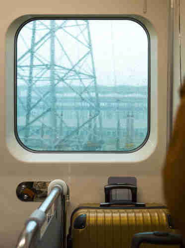 在捷运上, 一个金色的旅行箱靠窗放置. 窗外在下雨, 列车正好经过一个输电塔.