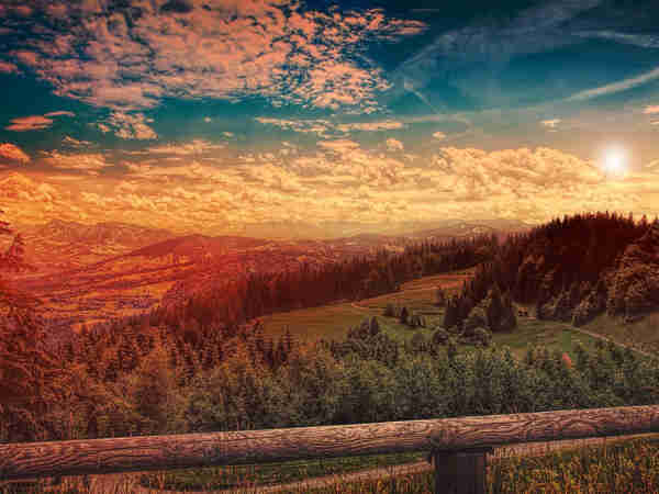 Warm coloured photo taken from a brown wooden balcony. In the foreground is a green hillside with many green pine trees on it. In the background are mountains with a red glow over them. The sky is yellow right above the horizon and deep blue above that, with many white clouds. 