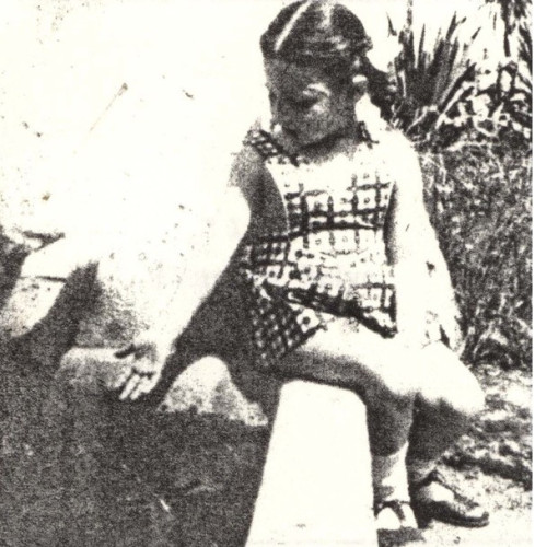 A young girl in a patterned dress is sitting outdoors on a sunlit day, focusing intently on something in their hands, surrounded by a natural, grassy environment.
