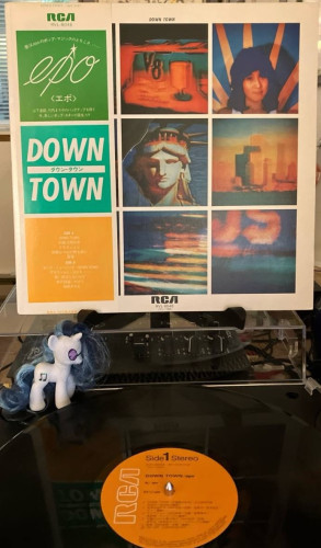 Record sleeve on a stand above a turntable. Sleeve features blurry photographs of America from a television set, including V8, a young woman, Statue of Liberty, New York City, and a flag.  The obi has RCA logo, "epo" in script and "DOWN TOWN" in block letters.

Below the turntable is a record with an orange label, with RCA logo visible.