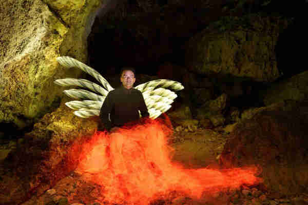 A person with illuminated wings and glowing effect around the lower body sitting in a rocky cave environment.
