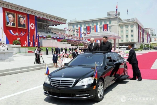 Putin and Kim sticking out of roof window of a luxury Mercedes limousine driving through a decorated square in Pyongyang 