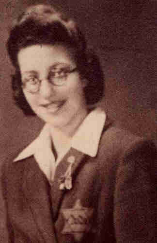 Portrait photograph of a young woman wearing a jacket and white shirt with a very wide collar. A Jewish star with the inscription 'Jude' is visible on the chest. The woman has a very high fringe. She is wearing glasses with round frames. She is smiling.