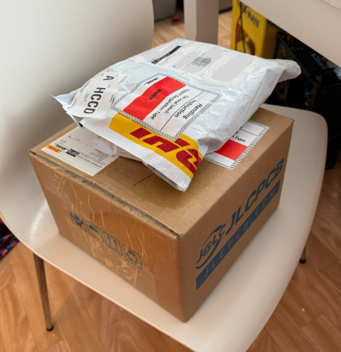 A large envelope and a box shipped by DHL stacked on top of one another on a chair. The lower box has JLCPCB printed on it in large blue letters.