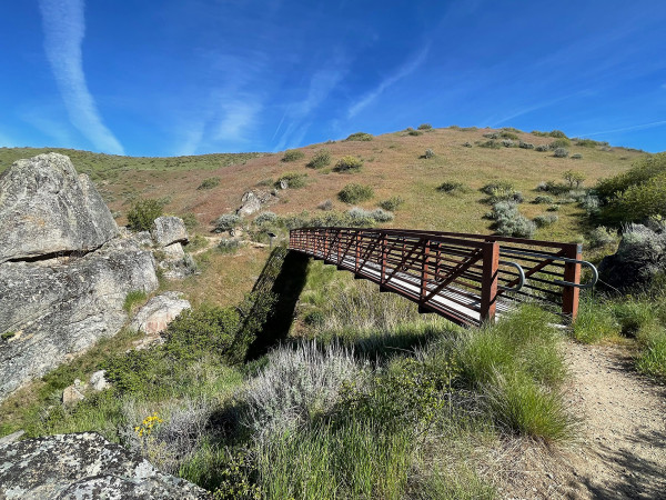 A foot bridge with iron posts and rails spans a gully. There are large granite rock outcroppings on the downhill side. A rolling hill dotted with sagebrush and rabbitbrush is above it. The sky is cloudless and has fading remnants of vapor contrails from airplanes.