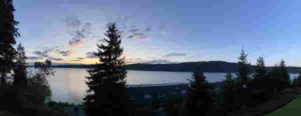 A panoramic view with a few evergreen trees in the front, the water of the Puget Sound, and pre-sunrise sky that's blue to the right and starting to turn yellow in the left where the sun is about to come up. 
