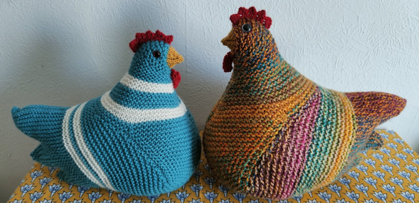 Two knitted chickens, one a solid teal and white, the other a mixture of all kinds of colours, sat face to face. The multicoloured chicken is bigger again than the teal one.