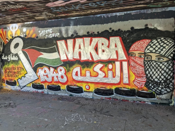 A graffiti mural showing the word "NAKBA" in large letters with the year 1948, an image of a flag, and two faces covered in keffiyehs. Text in Arabic and English references a generational struggle.