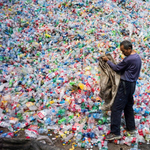 A man empties a canvas bag full of empty plastic bottles onto a gigantic mound of empty plastic bottles.