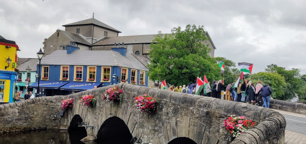 View of an arched stone bridge with flower baskets and colourful buildings & trees in the background. On the bridge people holding Palestinian flags