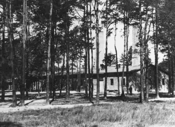 A long building behind trees. On the right - chimneys are visible. Crematorium V at Auschwitz II-Birkenau.