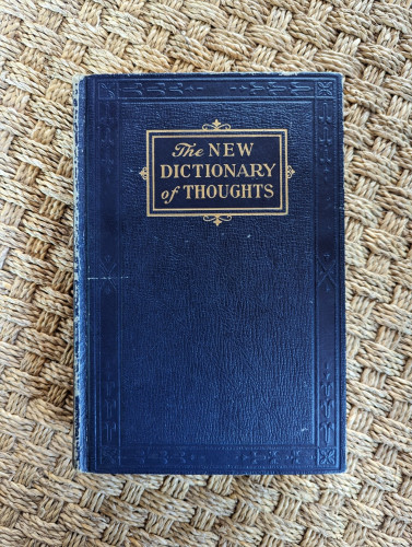 The front cover of an old book, titled The New Dictionary of Thoughts. The book is a royal blue color with the title in gold leaf lettering. There is some textured embellishment around the edges of the book, and the main center of the book is textured to look like leather. The book was printed in the 1960s or so. It's laying on a woven mat.