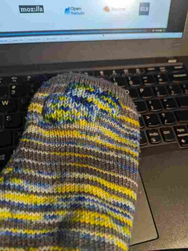A sock with duplicate stitch covering a thin spot in the sole. It is sitting on a laptop that is displaying the end of a FOSSDEM 24 (open source conference) video so there are a few logos from the conference's sponsors at the top of the photo. 
