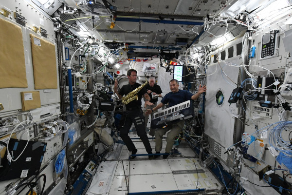 Three astronauts are floating in a space station filled with various scientific equipment, cables, and control panels. They are holding musical instruments: one has a saxophone, another has a keyboard, and the third has what appears to be a small drum or tambourine. The astronauts are in a playful pose, enjoying a moment of musical activity in the microgravity environment on World Music Day.