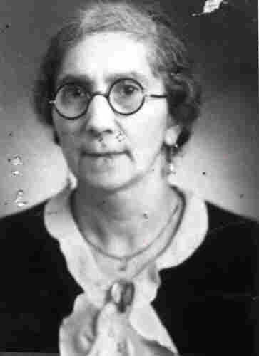 An ID portrait photograph of a mature woman. She has round, thin-framed glasses. She has a chain around her neck and a brooch on her collar.